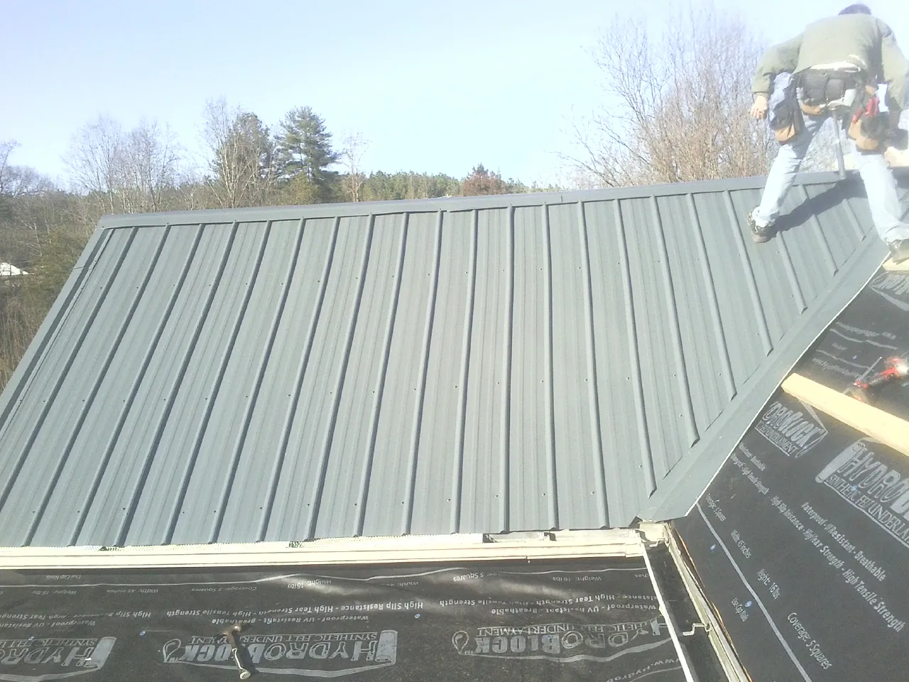 Crew installing dark green standing seam panels over synthetic underlayment in Otego