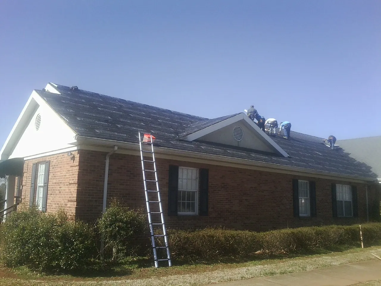 Crew tearing shingles off a brick ranch roof with a ladder set — Metal Roof Repair work by our crew in Otego