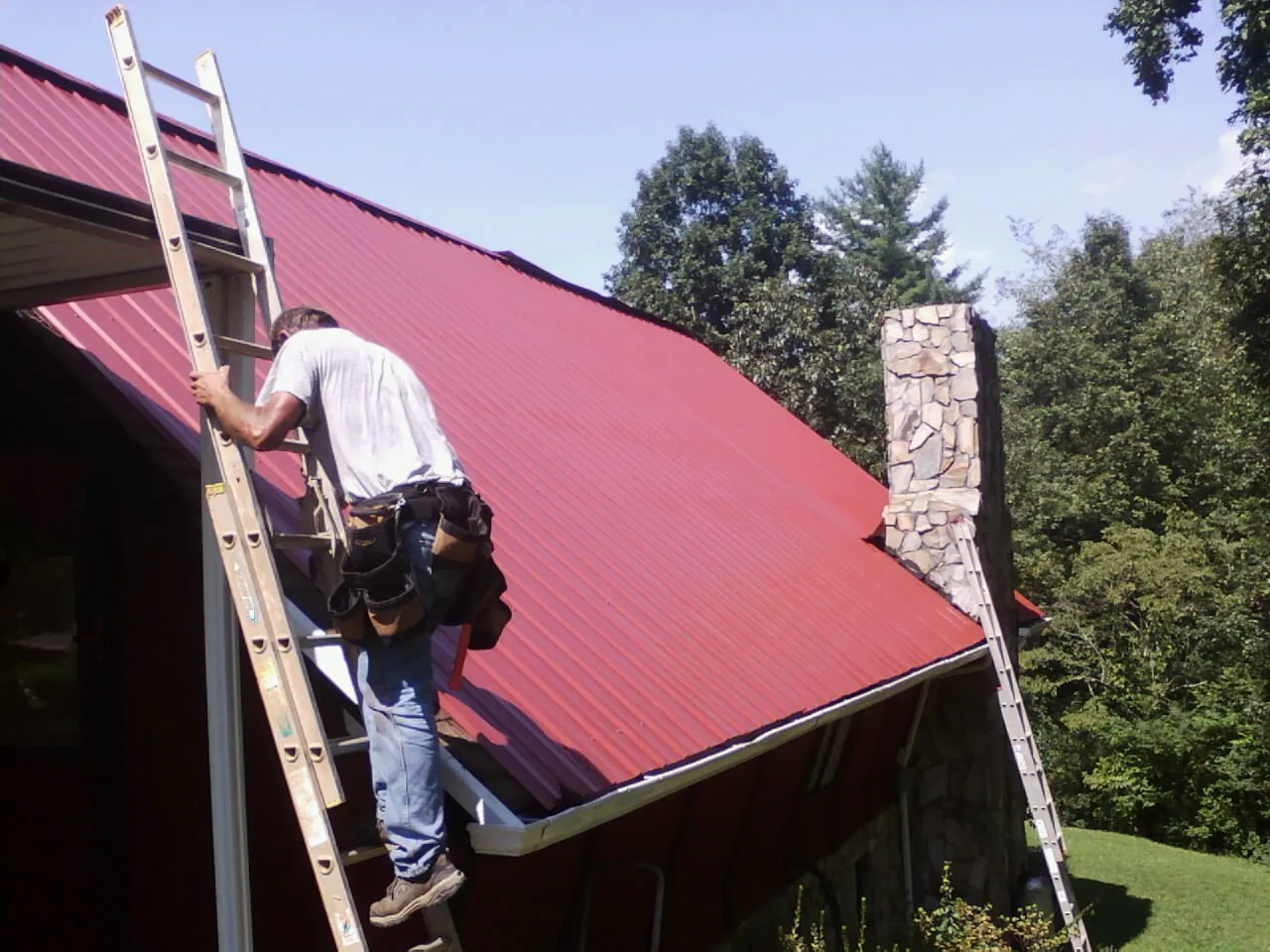 Roofer on a ladder beside a red metal roof and stone chimney — Soffit & Fascia Repair by Roofing Brothers in Otego